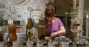 "Donna al lavoro in un antico laboratorio di erboristeria, con strumenti di distillazione, erbe essiccate e contenitori di vetro." Tisane autunnali