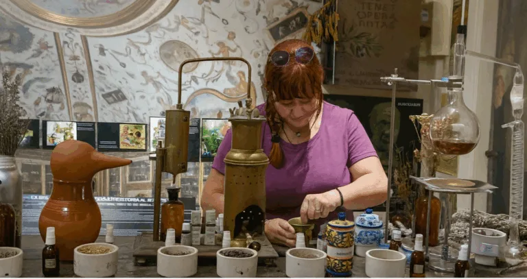 "Donna al lavoro in un antico laboratorio di erboristeria, con strumenti di distillazione, erbe essiccate e contenitori di vetro." Tisane autunnali